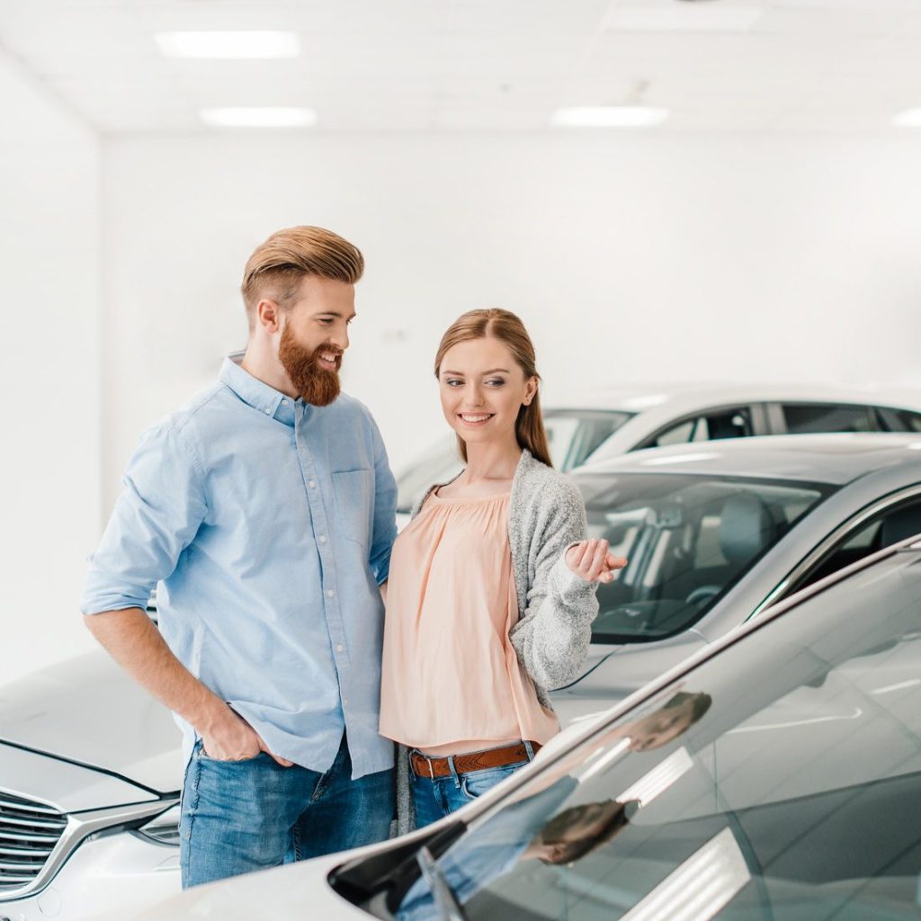 happy-couple-choosing-car-in-dealership-salon-woman-pointing-on-car-e1616394658847.jpg